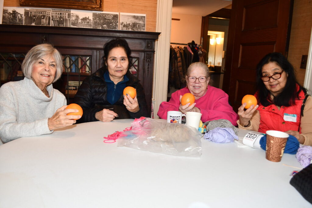 Four women sit around a table indoors, each holding an orange and smiling or looking toward the camera. Yarn, cups, and craft supplies are spread across the table, suggesting a casual group activity or social gathering in a cozy room with wooden furniture and framed photos on the wall.