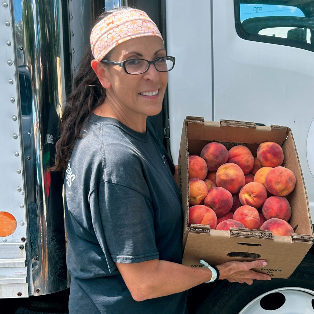 A woman wearing glasses and a patterned headband smiles while holding a cardboard box filled with fresh peaches beside a delivery truck.
