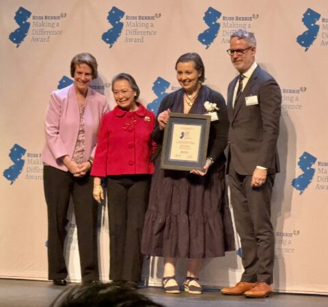 Four adults stand on a stage in front of a “Russ Berrie Making a Difference Award” backdrop, smiling for a photo. One woman at center-right holds a framed award certificate while wearing a dark dress and corsage; beside her, an older woman in a bright red jacket stands close. On the left, a woman in a light pink blazer smiles with hands clasped, and on the right, a man in a gray suit and glasses stands with hands at his sides.