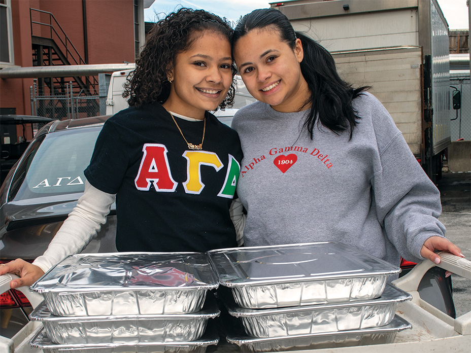 **Alt Text:** Two smiling young women stand close together behind a utility cart loaded with several large, foil-covered catering trays. The woman on the left has curly hair and wears a black t-shirt with the Greek letters "ΑΓΔ" (Alpha Gamma Delta). The woman on the right has straight hair pulled back and wears a grey sweatshirt featuring a red heart with "1904" and the words "Alpha Gamma Delta" printed on it.