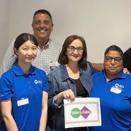 A group of seven diverse people standing together against a plain grey wall, smiling for a group photo. Several members wear blue polo shirts with the BD corporate logo, while one man wears a black BD polo shirt and gives a thumbs-up. A woman in a denim jacket stands near the center, holding up a printed sign displaying the green and purple "Table to Table" logo.