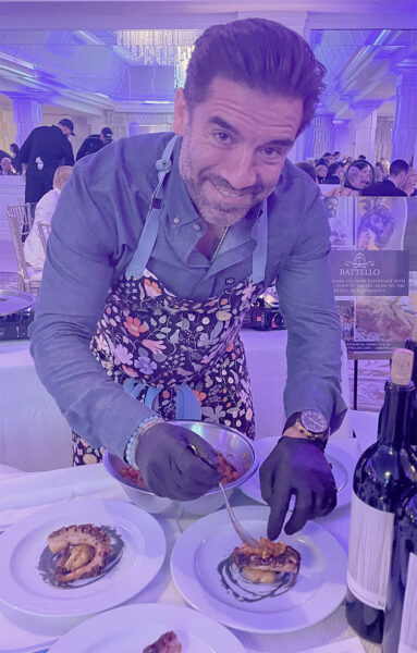 Chef Ryan DePersio with a beard, wearing a blue button-down shirt and a floral patterned apron, leans over a table to plate gourmet dishes at Table to Table's annual Chefs Gala. Wearing black food-preparation gloves, he uses a spoon to add a topping to a cooked octopus tentacle served on a white plate. The background shows a brightly lit, purple-hued ballroom with other guests, and a promotional sign for "Battello" is visible on the right.