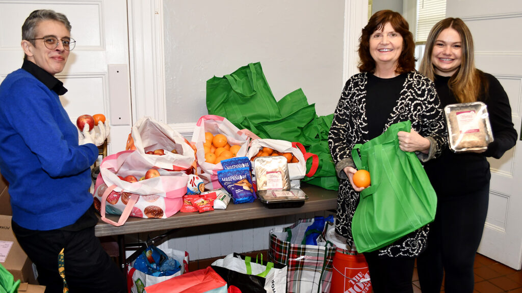 Three people smiling indoors next to a table overflowing with bags of groceries, including fresh fruit like apples and oranges, as well as packaged snacks, representing a food distribution or donation effort.