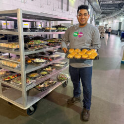 A man standing in a spacious indoor service corridor wears a long-sleeved gray shirt featuring the colorful "Table to Table" logo, dark jeans, and brown boots. He smiles while holding a black tray packed with small sandwich sliders wrapped in plastic wrap. Beside him on the left is a tall, multi-tiered metal utility cart on wheels, stacked with various plastic-wrapped food trays containing sandwiches, salads, and fruit platters.
