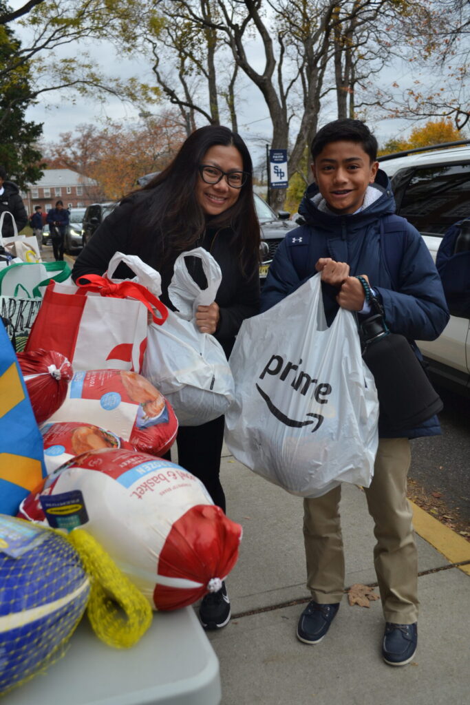 A smiling woman with glasses and a young boy holding a white Amazon Prime bag stand outdoors on a sidewalk during a Thanksgiving food drive. In the foreground, a table is filled with wrapped frozen turkeys and reusable grocery bags.