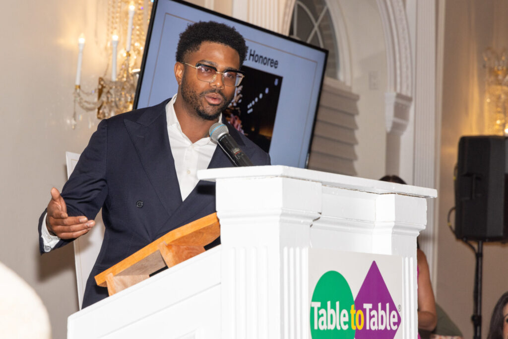 Chef Kwame Onwuachi wearing glasses and a dark suit standing at a white lectern featuring the "Table to Table" logo, speaking into a microphone with his right hand extended. In the background is a large screen displaying the word "Honoree" and elegant indoor wall sconce lighting.