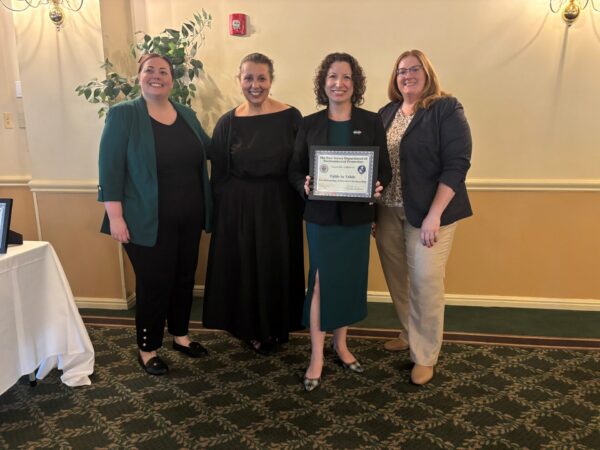 Four women stand side by side in an indoor event space, smiling for the camera. The woman second from the right holds a framed award certificate that reads "The New Jersey Department of Environmental Protection" and "Table to Table". The group is dressed in professional attire, standing in front of a light-colored wall with decorative moldings and green carpeted flooring.